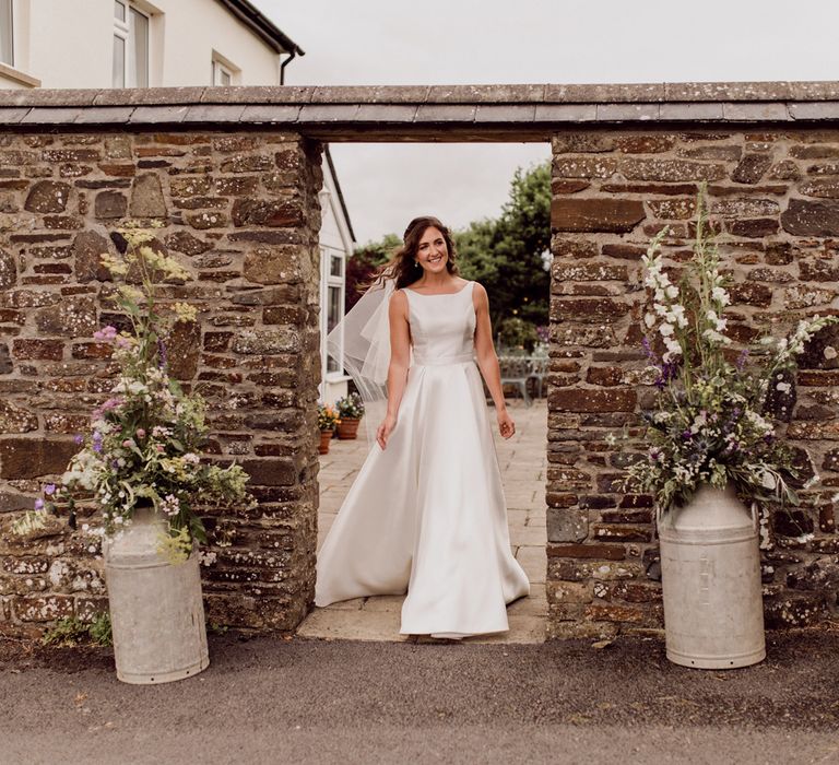 Bride in sleeveless satin Elbeth Gillis wedding dress and veil walks through doorway in brick wall before home farm wedding