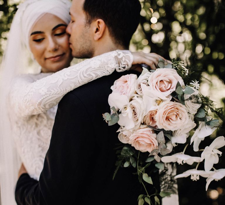 Groom kisses bride on the cheek who holds a pale pink rose and white orchid wedding bouquet