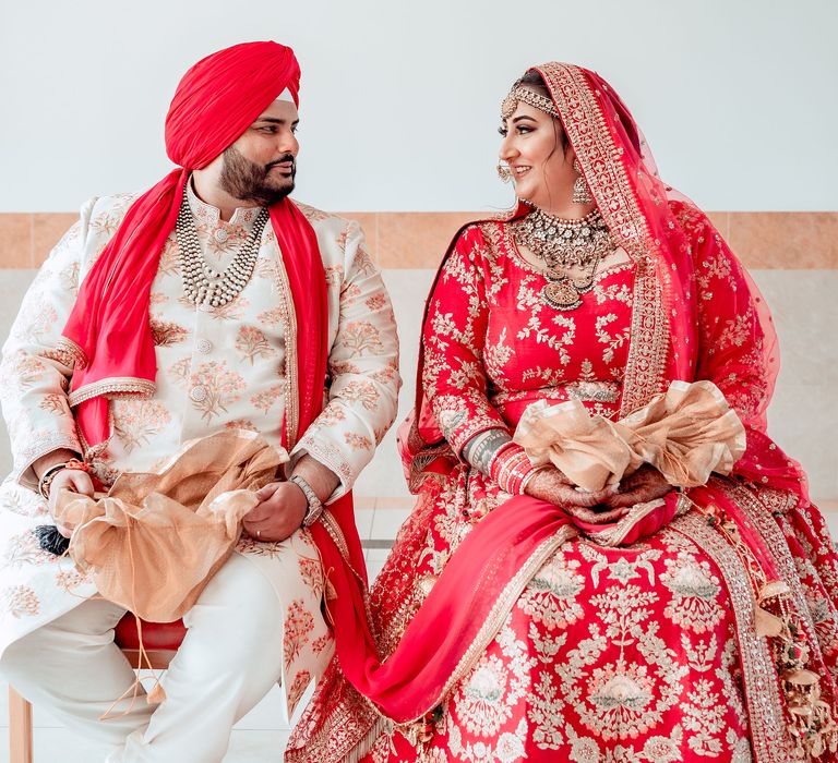 Bride & groom look lovingly at one another as bride wears traditional Lengha in red and gold, whilst groom wears cream coloured Kurta