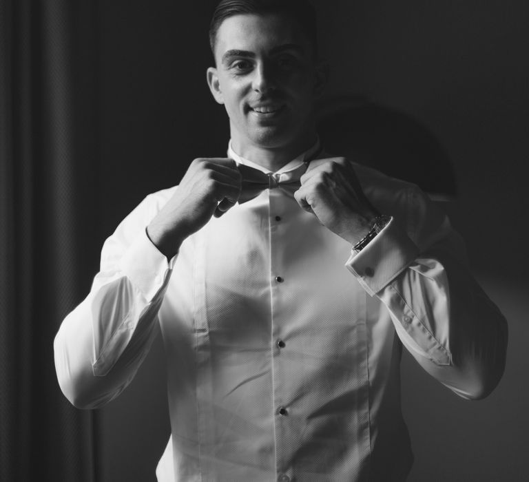 Groom does up his bow tie on the day of his wedding in black & white image