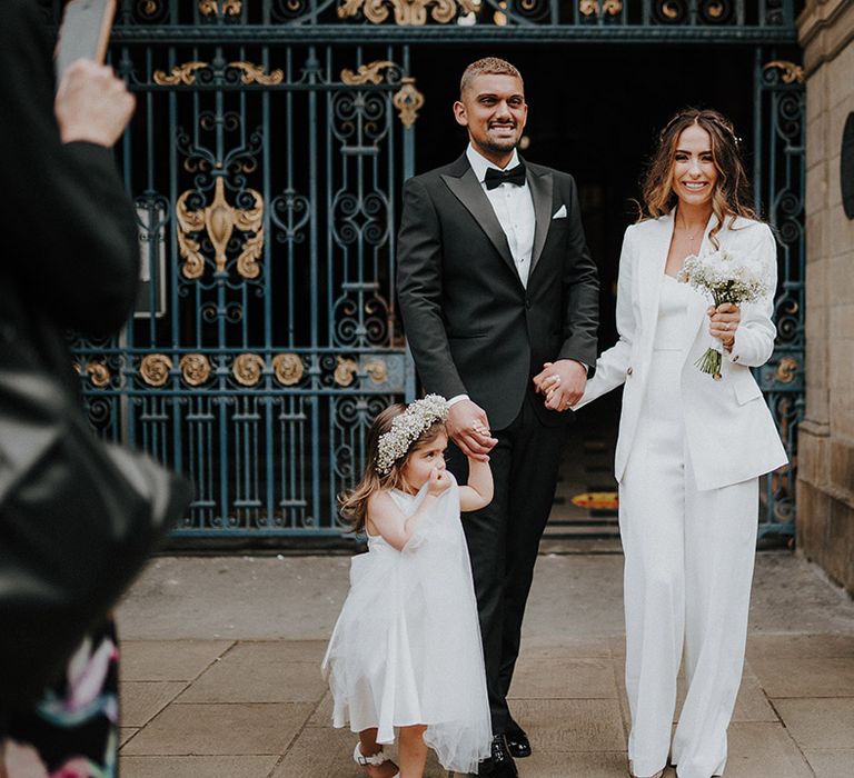 Family portrait with a groom in a tuxedo, bride in a suit and flower girl wearing a gypsophila flower crown 