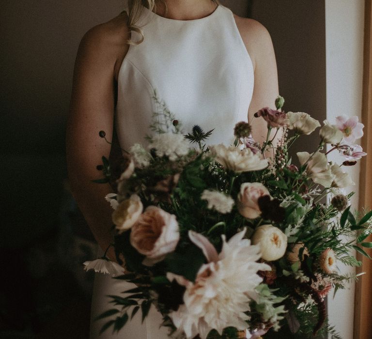 Blonde bride wears halter-neck wedding gown whilst looking down toward floral bouquet she is holding