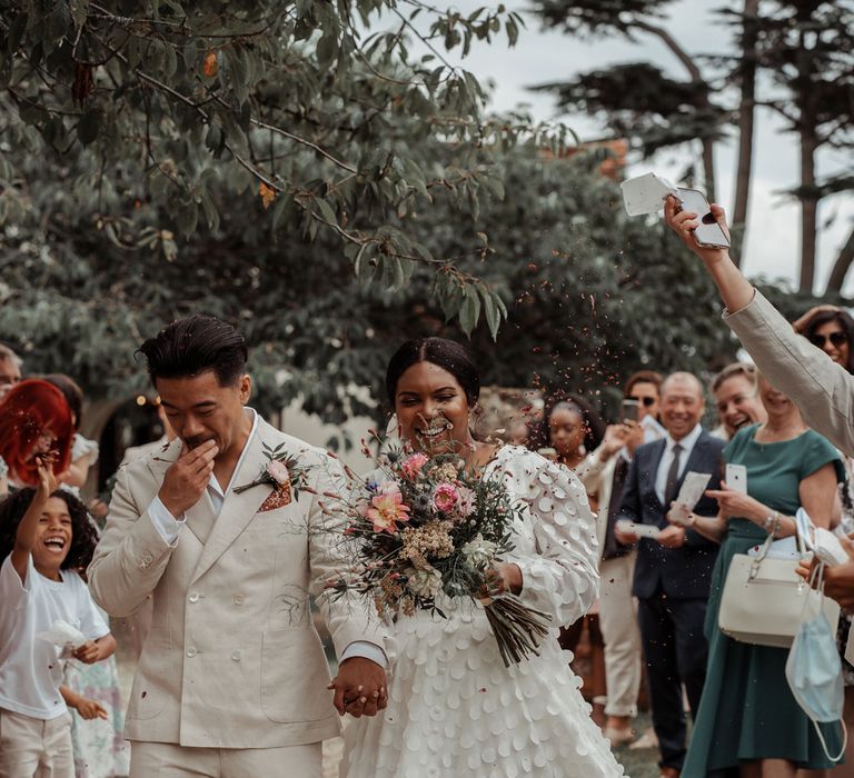 Smiling bride in white long mesh sleeve spotted Vagabond wedding dress holding mixed bridal bouquet walks holding hands with groom as guests throw confetti at Wasing Park wedding