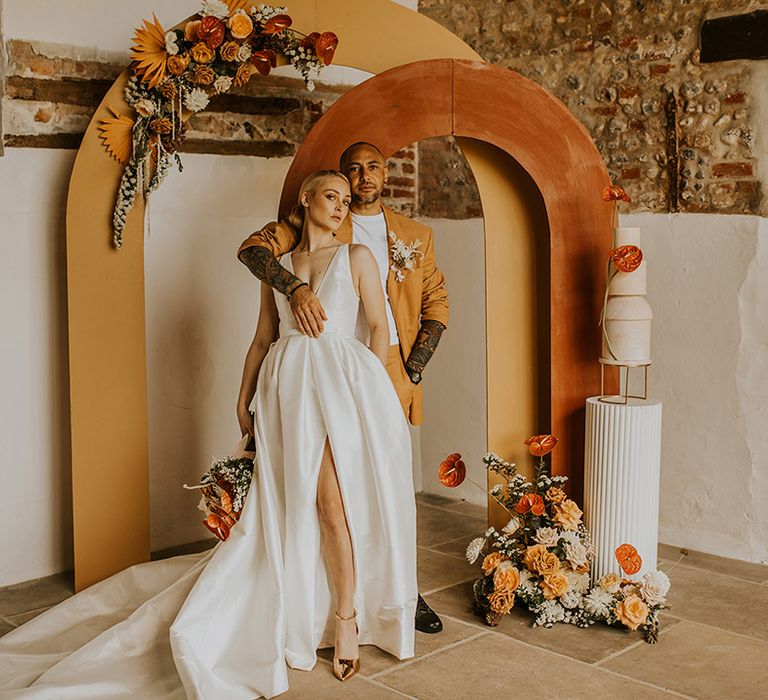 Stylish bride and groom in a Jesus Peiro wedding dress and mustard suit standing in front of yellow and orange arches decorated in autumnal flowers 