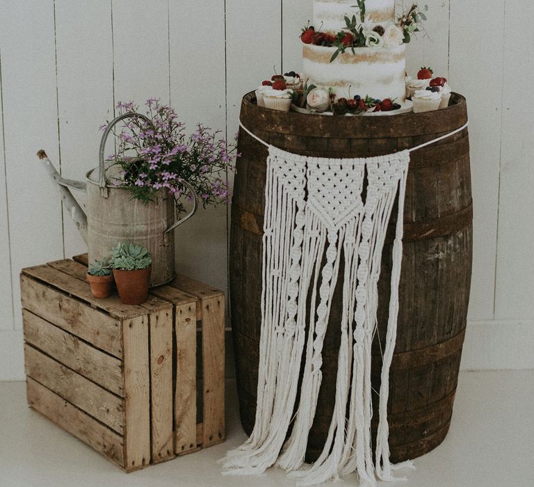 White three tier semi naked wedding cake with berry and foliage decor on wooden barrel with macrame hanging next to rustic wedding decor at Isle of Wight wedding