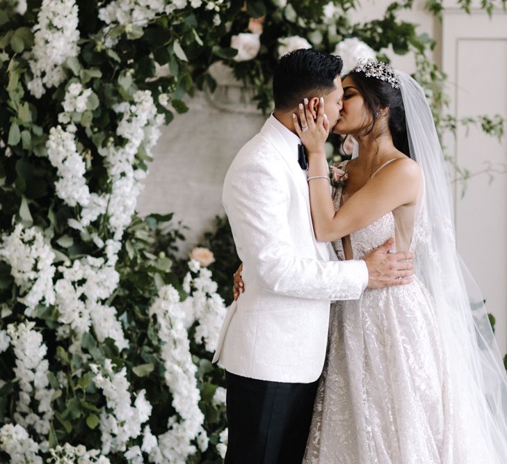 Bride in wedding dress and veil embraces and kisses groom in white suit jacket