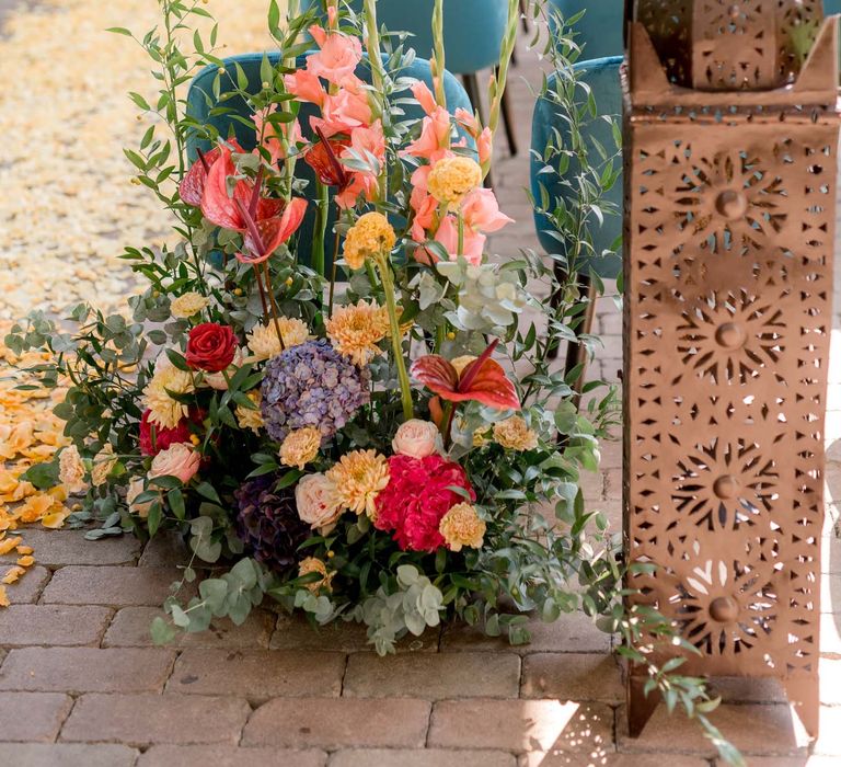 Beautiful and bright wedding florals sit at the end of aisle in front of emerald blue wedding chairs