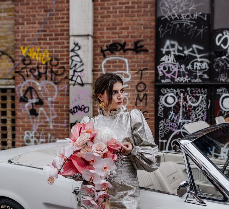 Bride holds bouquet of hydrangea, orchids and roses all full of the pink tones while she stands in front of vintage vehicle 