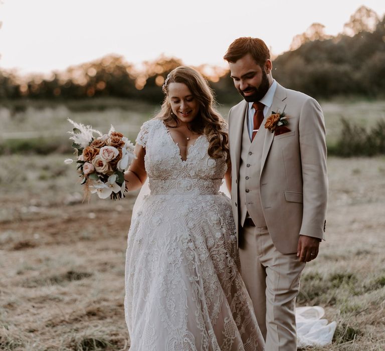 Birde in homemade lace wedding dress with train holding mixed rose bridal bouquet walks through field with groom in three piece beige suit, burgundy tie and floral buttonhole after DIY garden wedding ceremony