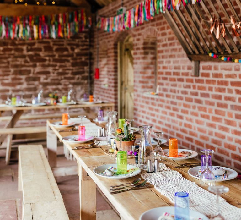 Long wooden wedding table with benches, colourful cups, macrame table runner and colourful streamers for rustic wedding breakfast at The Bridal Barn in Claverley