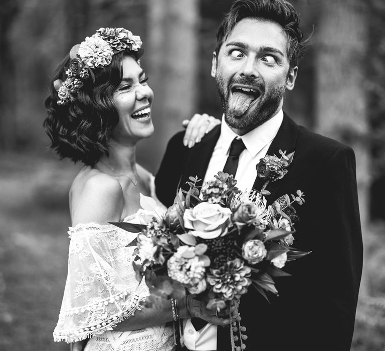 Bride in off the shoulder Grace Loves Lace wedding dress and colourful flower crown holds colourful wedding bouquet as she stand with groom in black suit as he pulls a face 