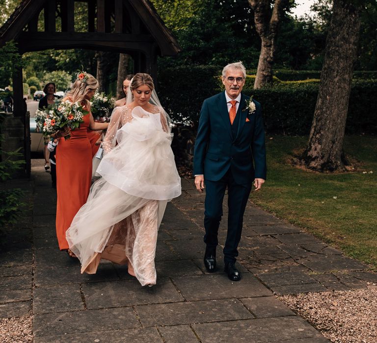 Bride in Julie Vino wedding dress and veil holds skirt as she walks to the church with father in blue three piece suit and bridesmaids in orange dresses before wedding ceremony