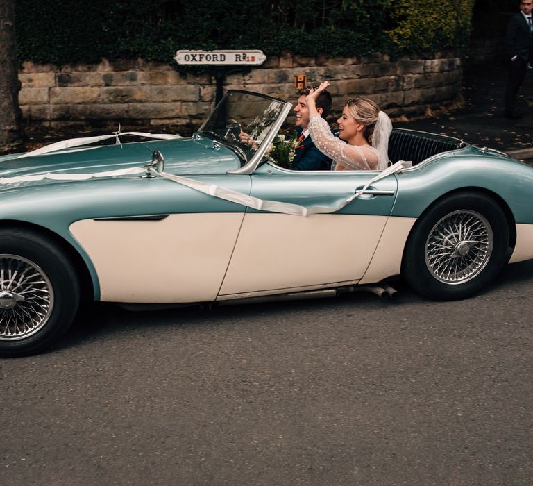 Bride in long mesh sleeve wedding dress and veil waves as groom in blue checked suit drives white and blue vintage sports car from church ceremony