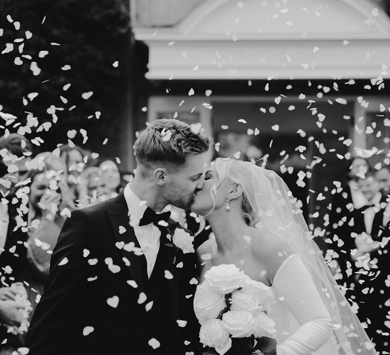 Black and white confetti portrait at Northbrook Park with bride in a strapless wedding dress with detachable sleeves kissing her groom in a tuxedo 