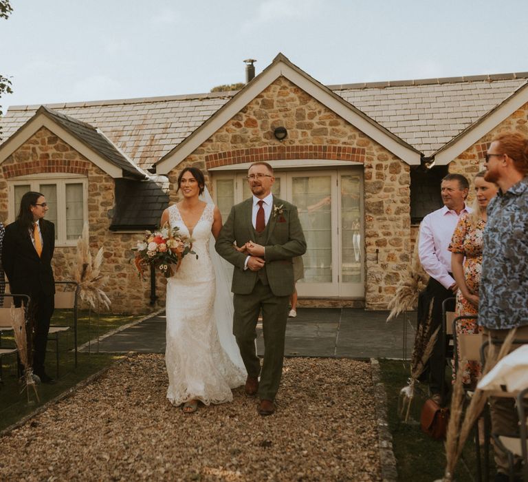 Bride in lace V neck wedding dress with train holding mixed bridal bouquet walks down outside aisle with man in green three piece suit and burgundy tie during outdoor wedding ceremony