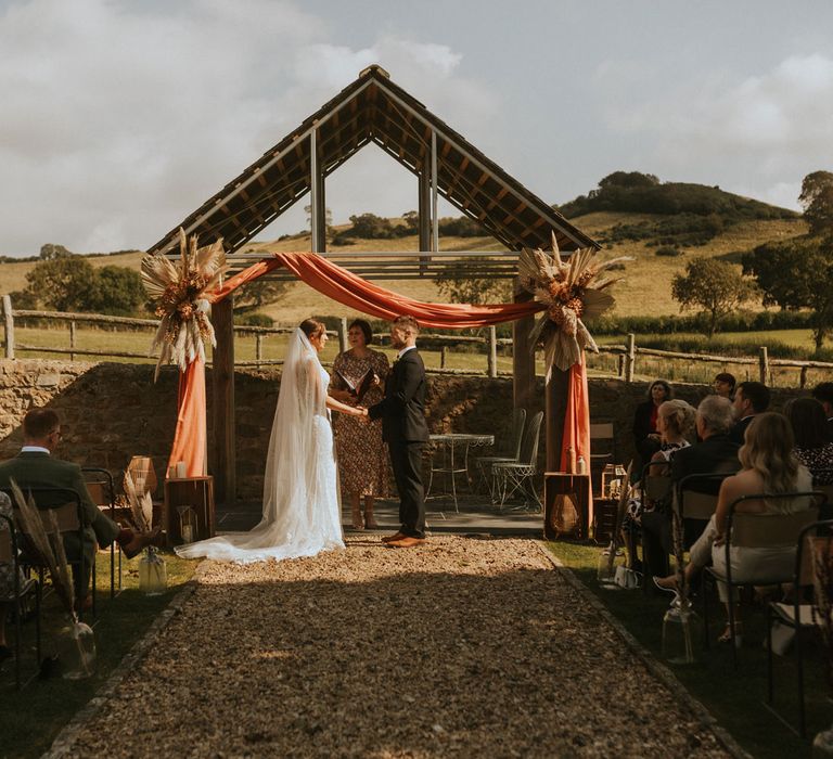 Bride in lace wedding dress with train and veil stands holding hands with groom in dark suit and brown shoes under rustic roof with orange draping for outdoor wedding ceremony