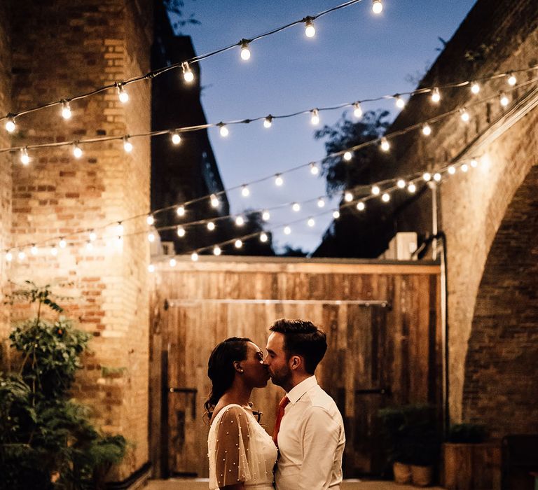 Black bride and groom kissing under a canopy of festoon lights at their 100 Barrington wedding 