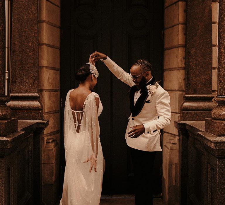 Black groom in a white tuxedo jacket twirling his bride in a fitted wedding dress with beaded Watteau train 