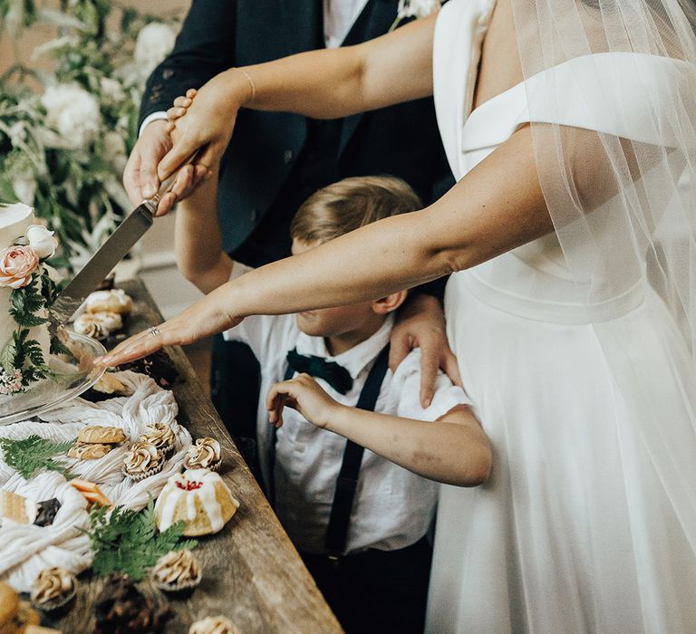 Bride in an off the shoulder wedding dress and veil cutting the cake with there groom and son 