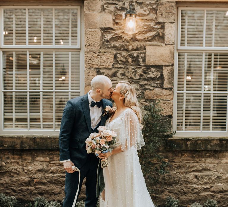 Bride in white pearl encrusted wedding dress with sleeves and train kisses groom in navy tweed suit with bow tie holding French bulldog on a lead