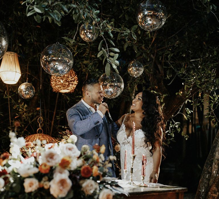 Groom in grey three-piece suit gives a speech with the bride standing next to him in lace floral wedding gown