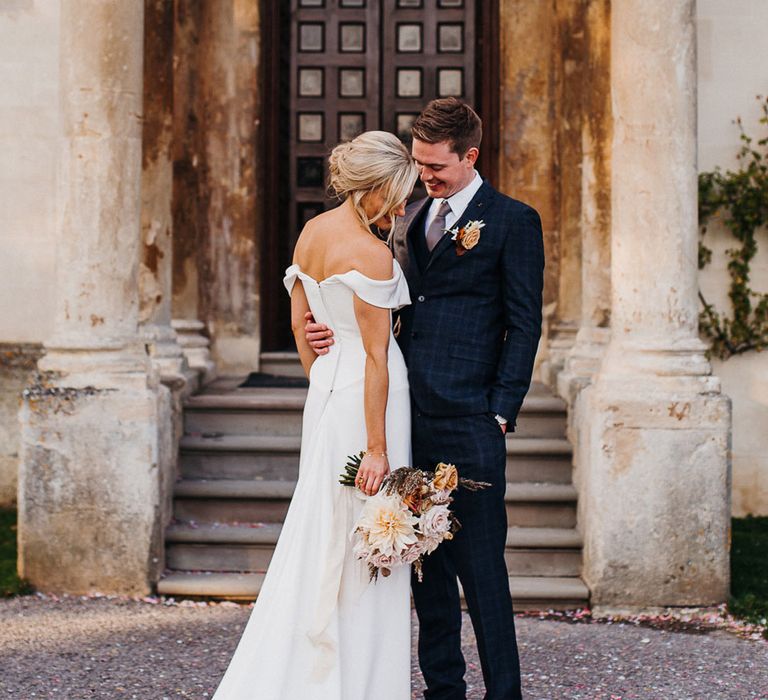 Bride holds pastel autumnal and neutral wedding bouquet as she poses with groom in blue suit 