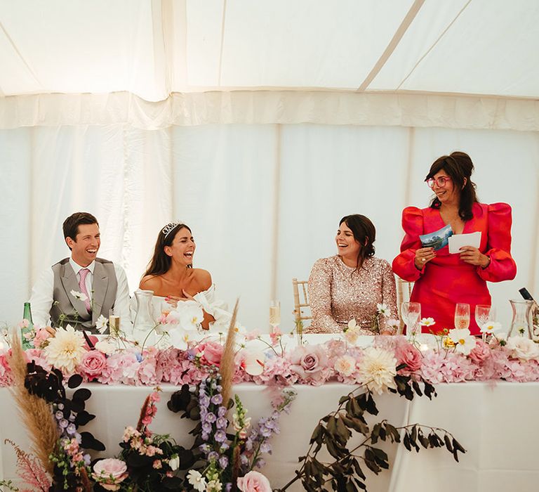 Groom in grey waistcoat and pink tie sits next to bride in off the shoulder dress as speeches are read out with pink, white and purple flower decorations