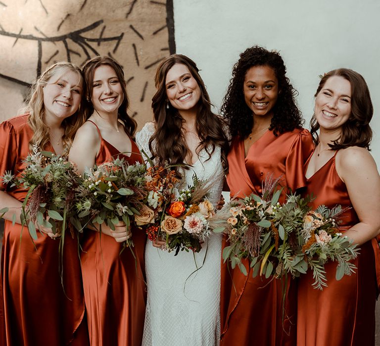 Bride in lace Maggie Sottero wedding dress smiles with bridesmaids in orange satin dresses with autumnal bouquets