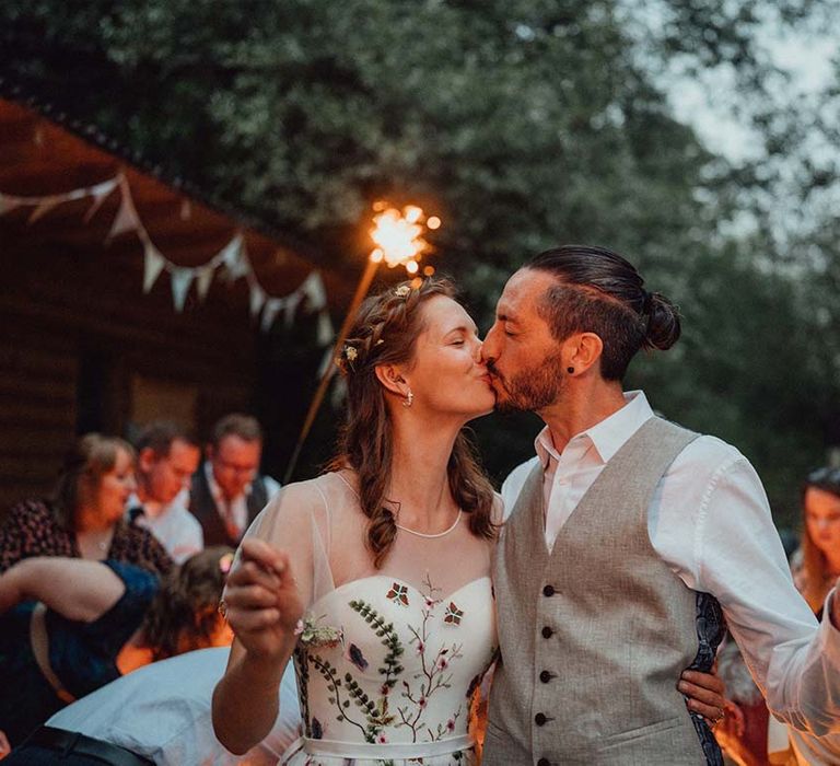 Groom in white shirt and grey waistcoat hugs with bride as they hold sparklers 