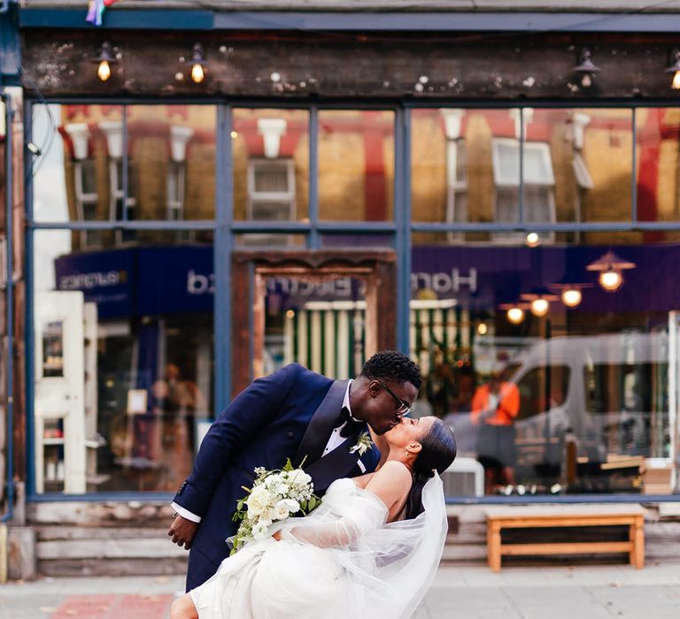 Groom leans the bride back as they kiss on crossing in black tie classic wedding outfits 