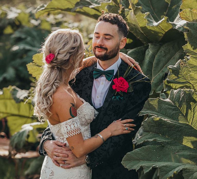 Bride in boho lace wedding dress with red flower in her hair hugs groom in black three piece patterned suit and red rose buttonhole 