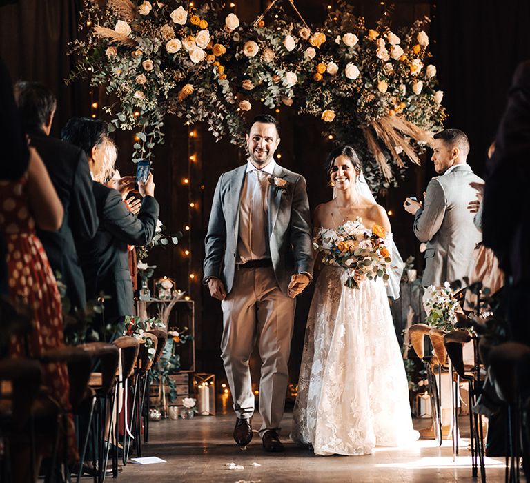 Bride and groom walk back down the aisle as a married couple with orange and white flower display with candles and dried flowers 