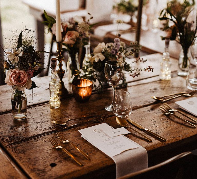 Wooden banquet tables with gold candle holders and cutlery with white menu card with wax seal with pink and white wedding flower arrangement