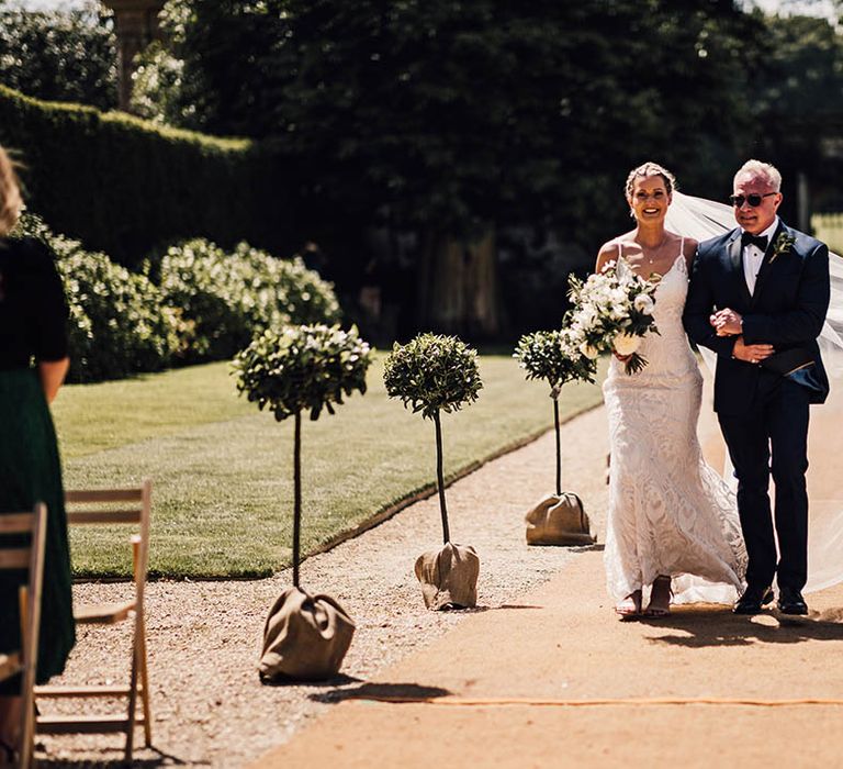 Father of the bride in blue tuxedo walks the bride down the aisle in lace wedding dress 