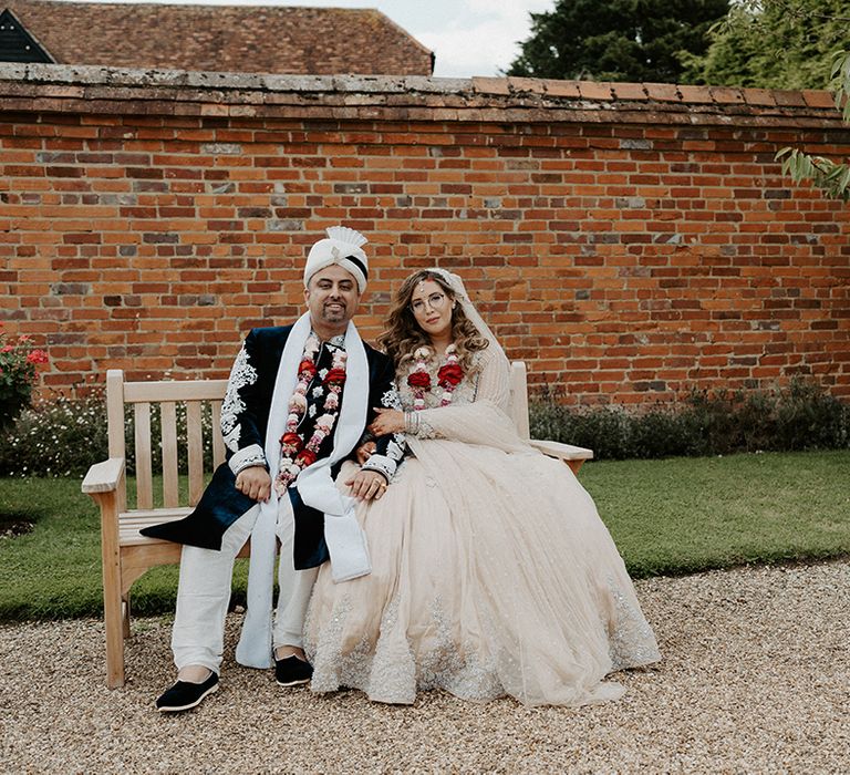 Bride & groom sit with one another wearing traditional floral garlands around their necks 