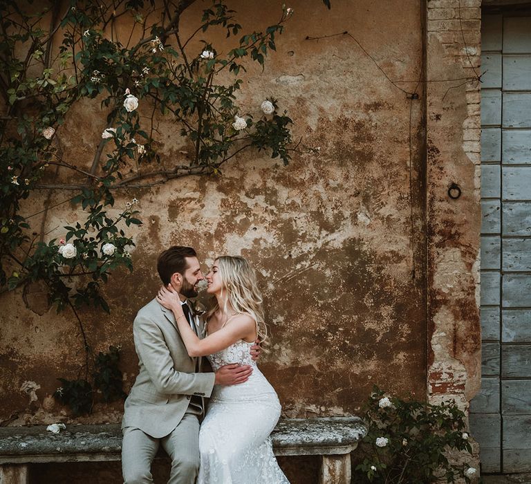 Bride & groom sit in front of rustic wall with vine climbing the wall as bride wears romantic lace wedding dress 