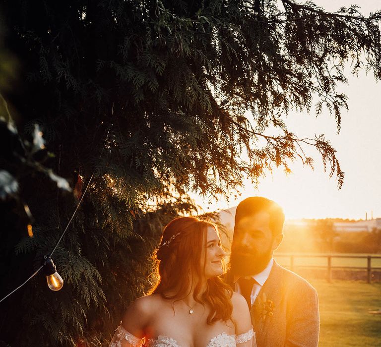 Bride in fitted boho lace wedding dress is hugged from behind by the groom in a grey suit and burgundy tie 