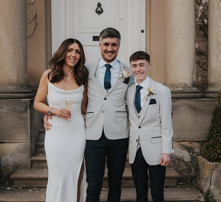 Bride and groom pose together with their ring bearer who wears a matching grey suit jacket with the groom 