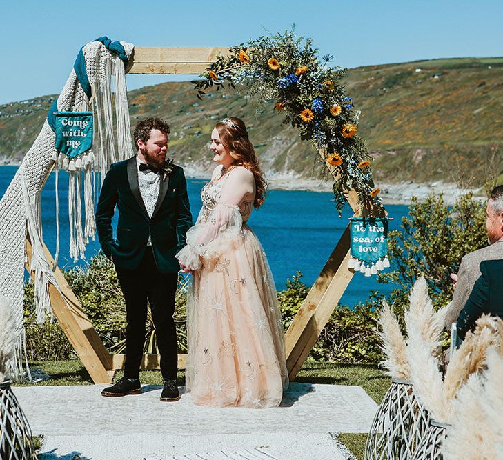 Bride & groom stand beneath DIY wooden arch finished with florals and wedding banners during ceremony outdoors on beach front in Cornwall