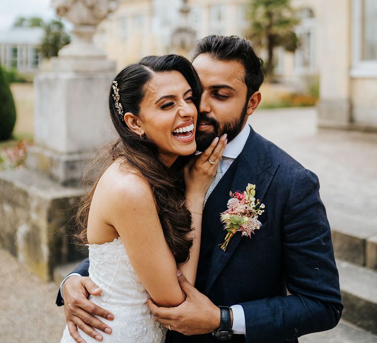 Groom wears three piece blue suit with floral buttonhole and stands beside his bride outdoors at Syon Park