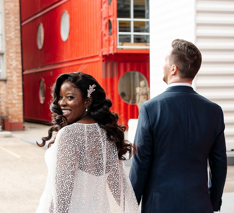 Black bride wears embellished hair accessory and tulle glitter wedding cape from Jesus Peiro as she walks with her groom 