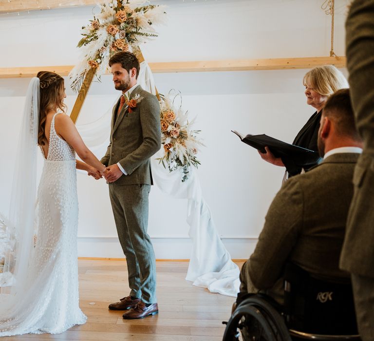 Bride in sleeveless Made With Love Bridal dress with white embellishments and groom in grey suit with burnt orange tie and pocket square holding hands at the alter standing by large wooden triangular arch