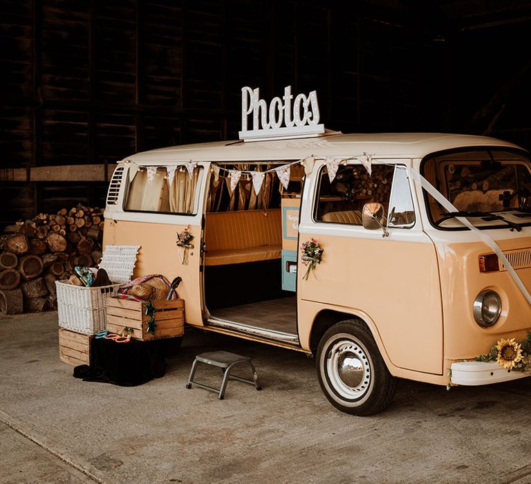 A pink VW camper photo booth decorated with white ribbon and sunflowers with a box of props for photos 