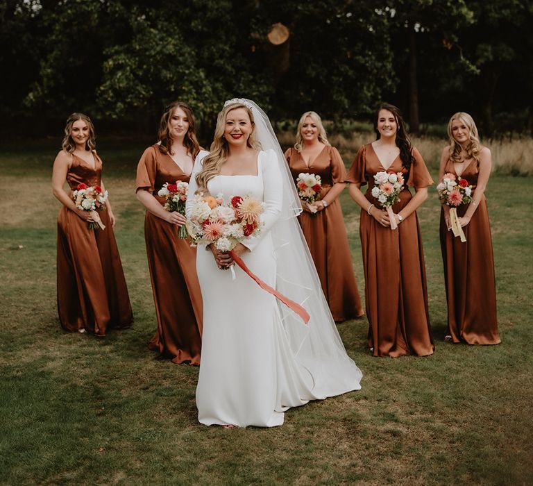 Bride in corset look bodice satin wedding dress and bridal party wearing burnt orange satin bridesmaid dresses holding red, pink, white and peach roses and dahlia wedding bouquets