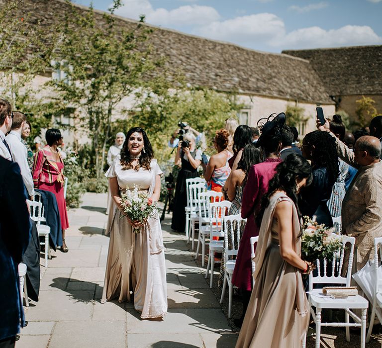 Bridesmaids in champagne bridesmaid dresses and Indian jewellery walk down the aisle at multicultural wedding