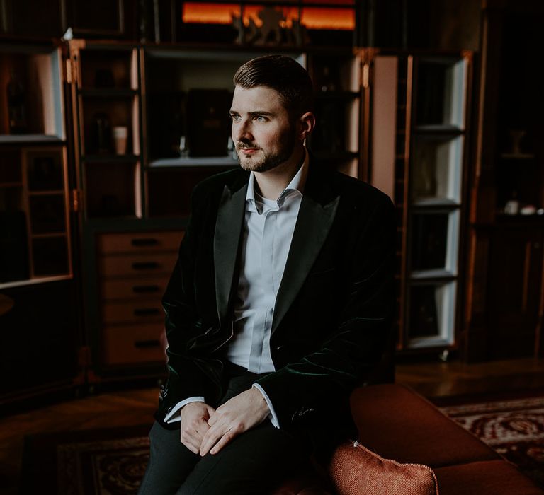 Groom in velvet tuxedo sitting gazing out the window on the day of his wedding 