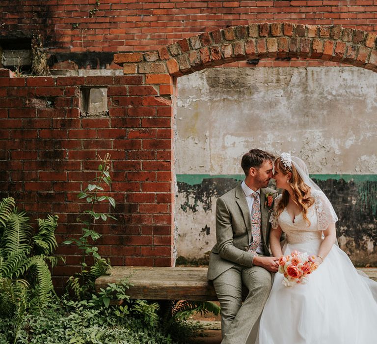 The bride and groom rest their foreheads against each other as they sit on a bench together 