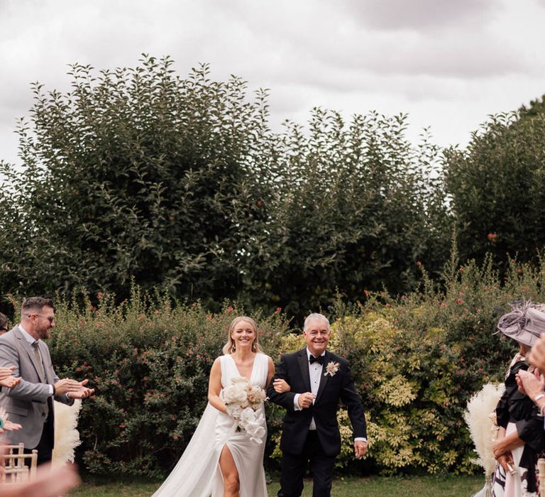 Father of the bride in black tie walks the bride in a plunging white wedding dress down the aisle 