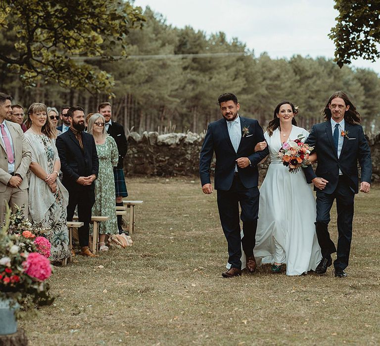 Bride in short sleeve v neck wedding dress with red lipstick walking down by two men in matching navy suits with different shades of blue ties