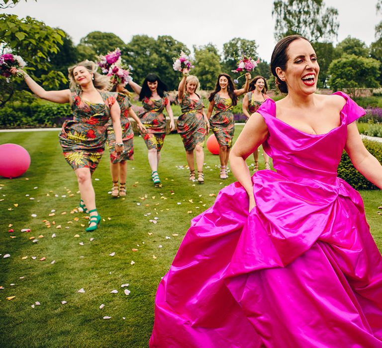 Bridesmaids in matching midi green colourful patterned Vivienne Westwood bridesmaid dresses with ruching, short sleeves and mismatched colour three strap shoes running after bride in off the shoulder front ruching pink wedding dress all holding pink floral bouquets with pink peonies, fuchsia roses, lilac sweet peas and green foliage